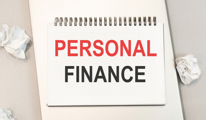 The word personal finance is written in a notebook that sits on a gray desktop along with a laptop.