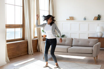 Happy young African American woman tenant relax rest dance alone in cozy light modern living room....