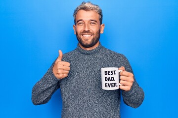 Handsome blond man drinking cup of coffee with best dad ever message over blue background smiling happy and positive, thumb up doing excellent and approval sign