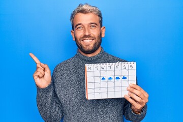 Young handsome blond man with beard holding weather calendar showing rainy week smiling happy...