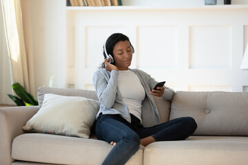 Obraz premium Happy calm young African American woman sit relax on sofa in living room listen music in headphones on cellphone. Relaxed biracial female rest on couch at home enjoy good quality sound in earphones.