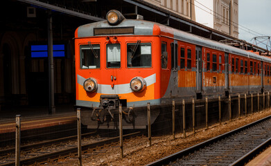 Obraz premium The train near the platform is waiting to be sent. Rail transport at the railway station. Red and orange train. Transportation is regular.