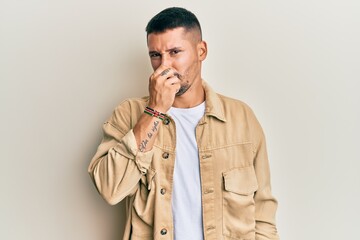 Handsome man with tattoos wearing casual brown denim jacket smelling something stinky and...