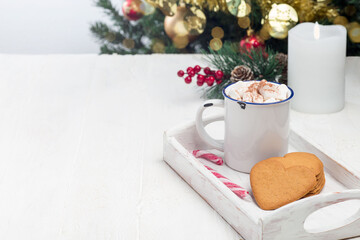 Fototapeta na wymiar Heart shaped gingerbread cookies with cup of hot chocolate and candy cane, on white wooden tray with christmas tree on background, horizontal, copy space