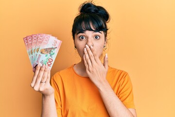 Young hispanic girl holding 100 new zealand dollars banknote covering mouth with hand, shocked and afraid for mistake. surprised expression