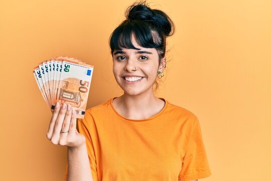 Young hispanic girl holding bunch of 50 euro banknotes looking positive and happy standing and smiling with a confident smile showing teeth