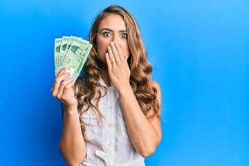 Young blonde girl holding 10000 south korean won banknotes covering mouth with hand, shocked and afraid for mistake. surprised expression