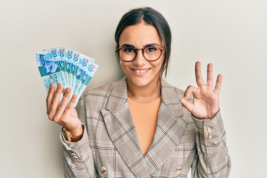 Young brunette woman holding hong kong 20 dollars banknotes doing ok sign with fingers, smiling friendly gesturing excellent symbol