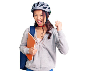 Young beautiful chinese girl wearing student backpack and bike helmet holding book screaming proud, celebrating victory and success very excited with raised arms