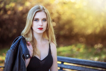 Attractive girl with a leather jacket on her shoulder is sitting on a bench in autumn park. Horizontally. 