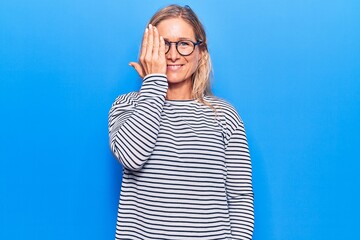 Obraz na płótnie Canvas Middle age caucasian blonde woman wearing casual striped sweater and glasses covering one eye with hand, confident smile on face and surprise emotion.