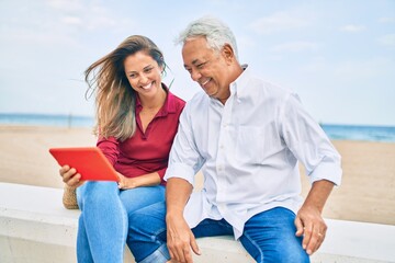 Middle age hispanic couple using touchpad sitting on the bench at the beach.