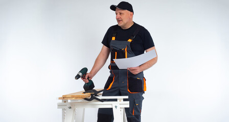 Carpenter with a cordless screwdriver in his hands. A man in work clothes holds drawings in his hands and communicates with a colleague backstage. Joiner in a gray jumpsuit and a baseball cap.