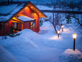 Christmas in the village. Cottage with garlands on the background of snow. Winter evening away from the city. A path in the snow leads to the house with garlands. The concept of home comfort.