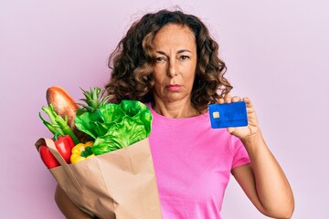 Middle age hispanic woman holding groceries and credit card depressed and worry for distress, crying angry and afraid. sad expression.