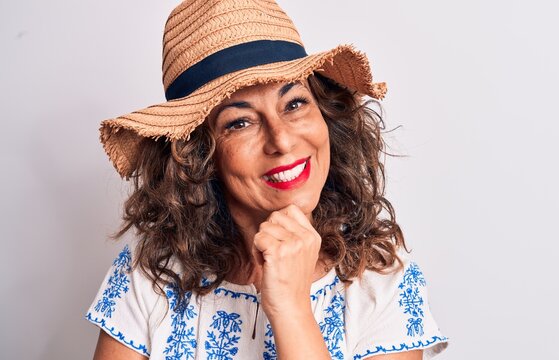 Middle age beautiful brunette woman on vacation wearing summer hat over white background smiling looking confident at the camera with crossed arms and hand on chin. Thinking positive.