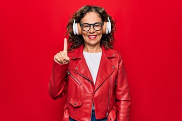 Middle age beautiful woman listening to music using headphones over red background smiling with an idea or question pointing finger up with happy face, number one