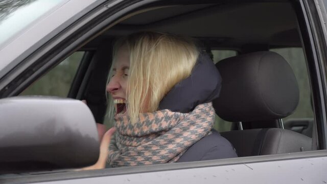 A young blonde gets angry and screams while waiting in traffic. She screams and slams her hands on the steering wheel, the car broke down, she is in the middle of the forest in nature.
