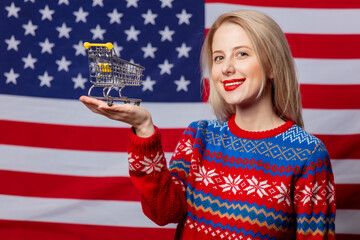 Beautiful blonde in Christmas sweater holds shopping cart on USA flag background