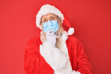 Old senior man wearing santa claus costume wearing safety mask smiling looking confident at the camera with crossed arms and hand on chin. thinking positive.