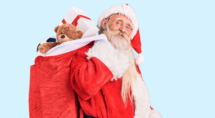 Old senior man with grey hair and long beard wearing santa claus costume holding bag with presents looking positive and happy standing and smiling with a confident smile showing teeth