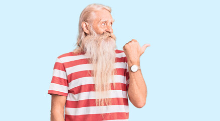 Old senior man with grey hair and long beard wearing striped tshirt smiling with happy face looking and pointing to the side with thumb up.