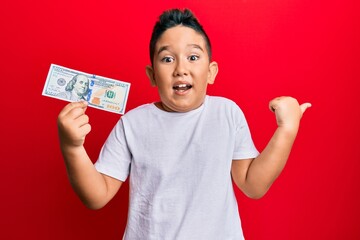 Little boy hispanic kid holding 100 dollars banknote pointing thumb up to the side smiling happy with open mouth