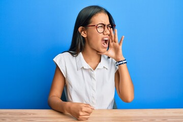 Beautiful hispanic woman wearing casual clothes sitting on the table shouting and screaming loud to side with hand on mouth. communication concept.