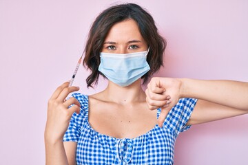Young beautiful woman wearing medical mask holding syringe with angry face, negative sign showing...