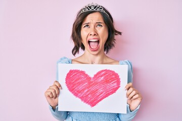 Young beautiful girl wearing princess crown holding heart draw angry and mad screaming frustrated and furious, shouting with anger looking up. © Krakenimages.com