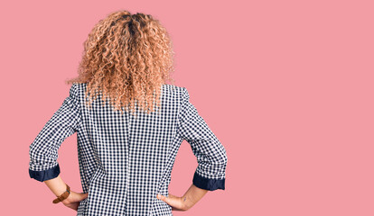 Young blonde woman with curly hair wearing business jacket and glasses standing backwards looking away with arms on body