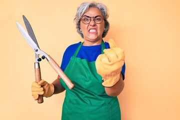 Senior hispanic woman wearing gardener apron and gloves holding shears annoyed and frustrated...