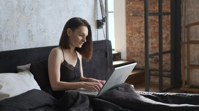 woman in nightie works at laptop while lying in bed