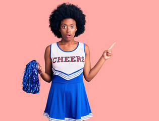 Young african american woman wearing cheerleader uniform holding pompom surprised pointing with...