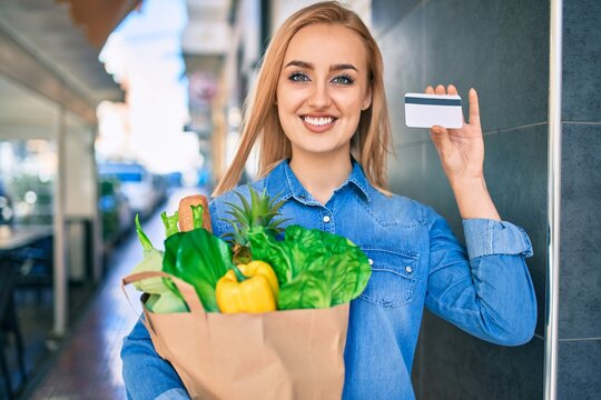 Young blonde girl smiling happy holding groceries paper bag and credit card standing at the city.