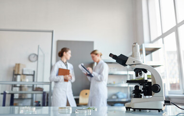Naklejka premium Portrait of two female scientists discussing research while standing in medical laboratory, focus on microscope in foreground, copy space
