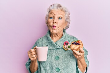 Senior grey-haired woman drinking a cup of coffee and eating bun making fish face with mouth and...