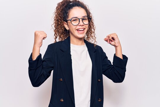 Beautiful kid girl with curly hair wearing business clothes and glasses screaming proud, celebrating victory and success very excited with raised arms