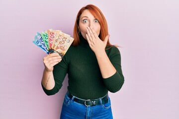 Beautiful redhead woman holding canadian dollars covering mouth with hand, shocked and afraid for...