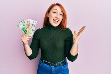 Beautiful redhead woman holding russian ruble banknotes celebrating achievement with happy smile and winner expression with raised hand