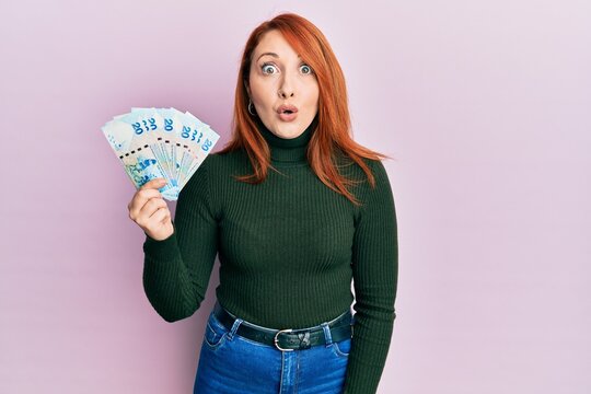 Beautiful redhead woman holding 20 hong kong dollars banknotes scared and amazed with open mouth for surprise, disbelief face