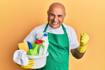 Mature middle east man wearing cleaner apron holding cleaning products screaming proud, celebrating victory and success very excited with raised arms