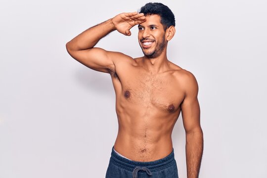 Young latin man standing shirtless very happy and smiling looking far away with hand over head. searching concept.