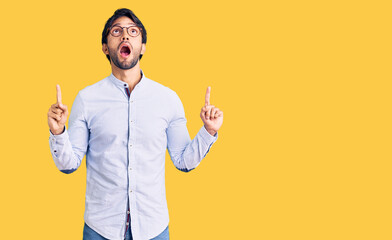 Handsome hispanic man wearing business shirt and glasses amazed and surprised looking up and pointing with fingers and raised arms.