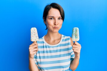 Young brunette woman with short hair holding two ice cream puffing cheeks with funny face. mouth inflated with air, catching air.