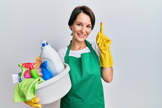 Young brunette woman with short hair wearing apron holding cleaning products pointing finger up with successful idea. exited and happy. number one.