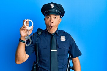 Handsome middle age mature man wearing police uniform holding metal handcuffs scared and amazed with open mouth for surprise, disbelief face