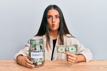 Beautiful hispanic woman holding charity jar with one dollar banknote making fish face with mouth and squinting eyes, crazy and comical.