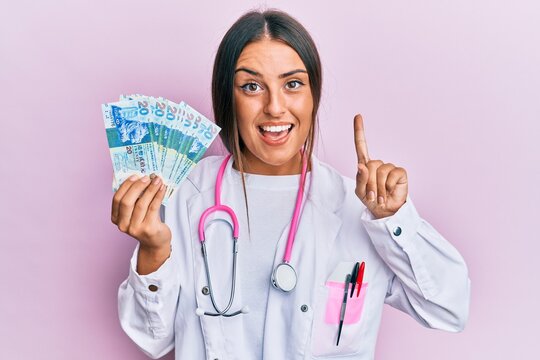 Beautiful hispanic woman wearing medical uniform holding 20 hong kong dollars smiling with an idea or question pointing finger with happy face, number one