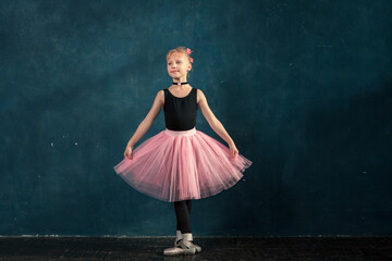 Caucasian little girl in pink tutu and pointe shoes training ballet position against wall in studio